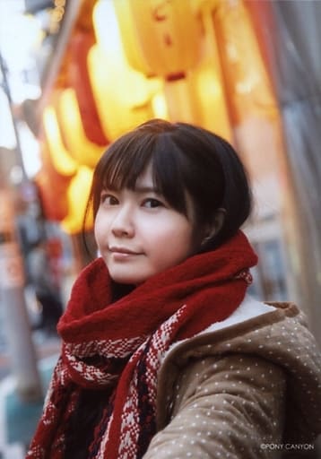 Taketatsu Ayana in a brown jacket and a red muffler, smiling with her left arm stretched outwards