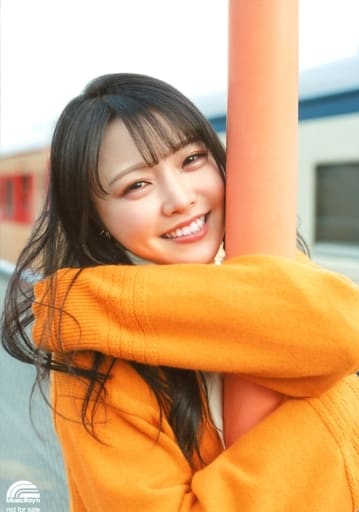 Asakura Momo in a orange sweater hugging on to an off-colour orange thin pole smiling back with a train in the background