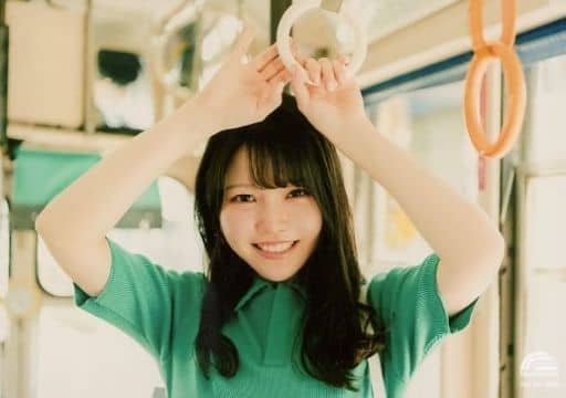 Asakura Momo smiling in a green blouse with a white sling handbag, holding on to a handrail in a train